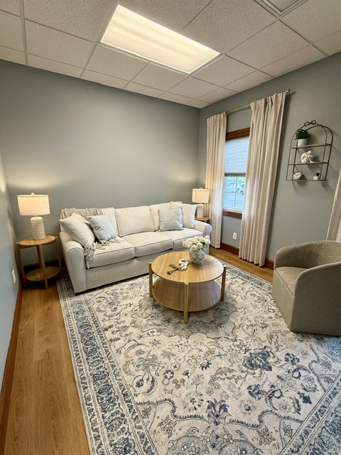 Anchor Within Counseling therapy room in Hamburg, NY with a soft cream sofa, accent chair, round coffee table with fresh flowers, and a patterned area rug in calming sage tones.