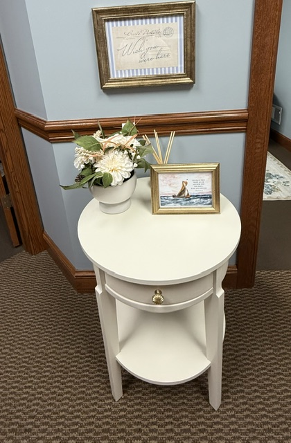 A warm entry detail with a round white side table, fresh flowers, and gently framed coastal artwork against blue walls and natural wood trim.